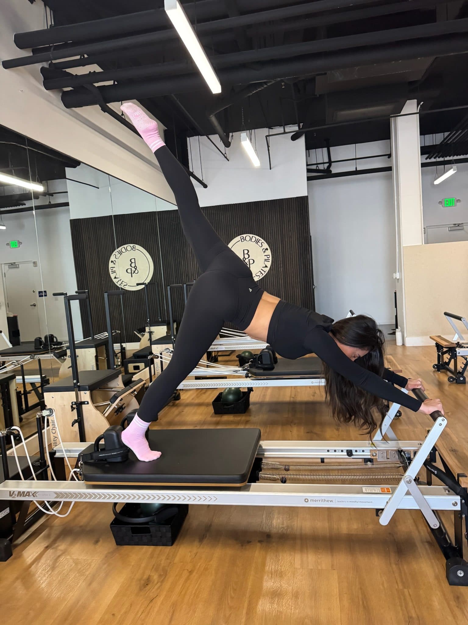 Standing split on the reformer in front of the logo wall at Bodies and Pilates in North Hollywood